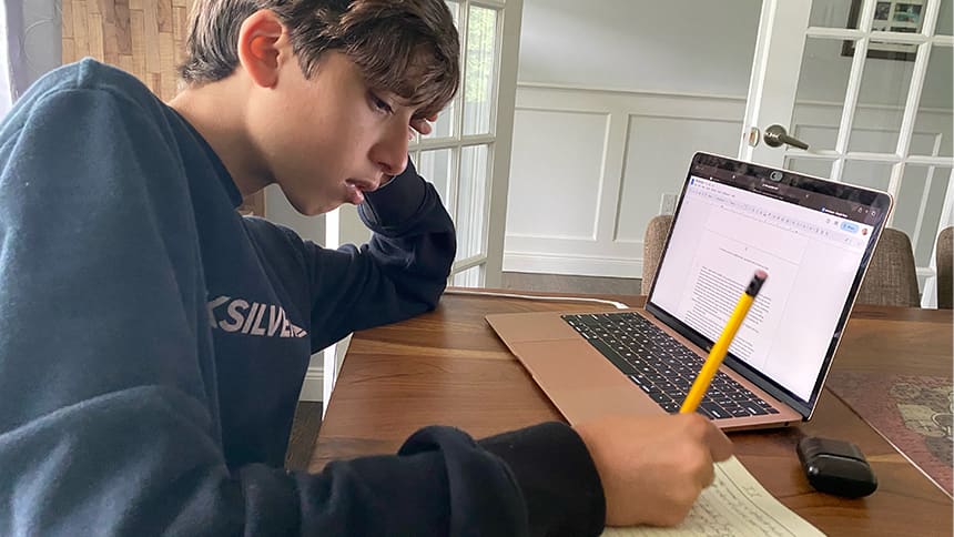 Taym sitting at a table in front of a laptop, writing in a notebook.