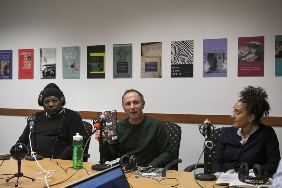 Three people seated at a table. The person in the middle is holding a book.