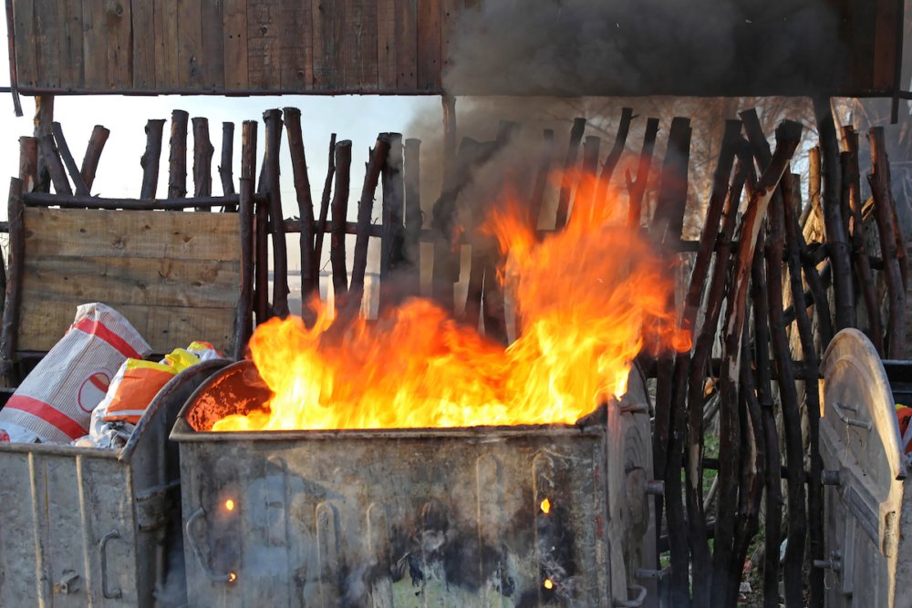 Image: flames and smoke erupt from a garbage dumpster.