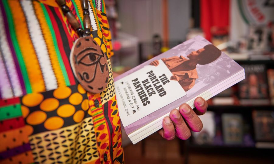 a close-up of a hand holding the book “The Portland Black Panthers” with bookshelves in the background